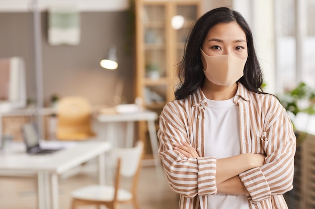 Taille portrait de jeune femme asiatique portant un masque et regardant un appareil photo en se tenant debout avec les bras croisés dans un bureau moderne, copiez l'espace