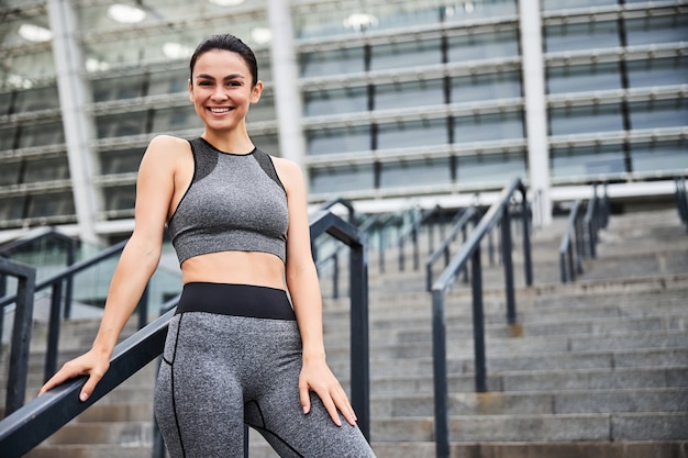 Taille portrait d'une jolie femme joyeuse en tenue de sport debout dans les escaliers menant au grand stade urbain