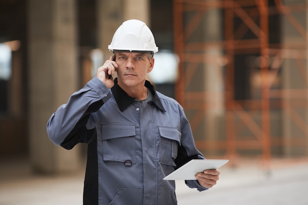 Taille portrait de travailleur d'âge mûr parlant par smartphone et debout sur un chantier de construction ou dans un atelier industriel,