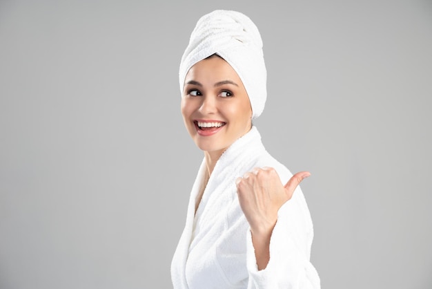 Taille vue portrait de la belle femme avec une serviette sur la tête, vêtue d'un peignoir blanc en détournant les yeux et pointant sur le fond gris du studio. Concept de photo de beauté