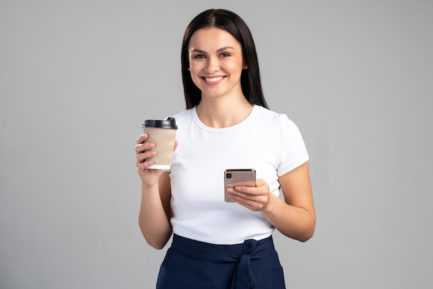 Taille vue portrait de la belle femme tenant une tasse de délicieux café délicieux dans les mains et smartphone isolé sur fond gris