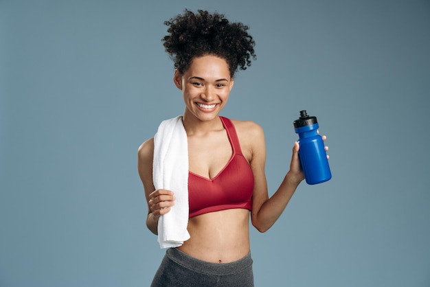 Taille vue portrait de la femme multiraciale en bonne santé buvant de l'eau après avoir fait de l'exercice tout en tenant une serviette et en regardant la caméra