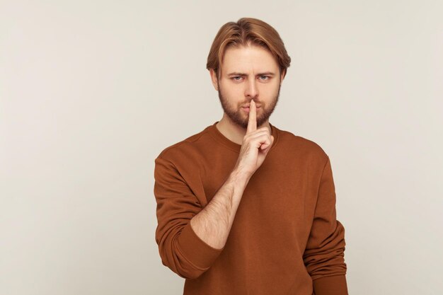 Tais-toi, tais-toi. Portrait d'un homme mystérieux avec barbe portant un sweat-shirt montrant un geste de silence avec le doigt sur les lèvres