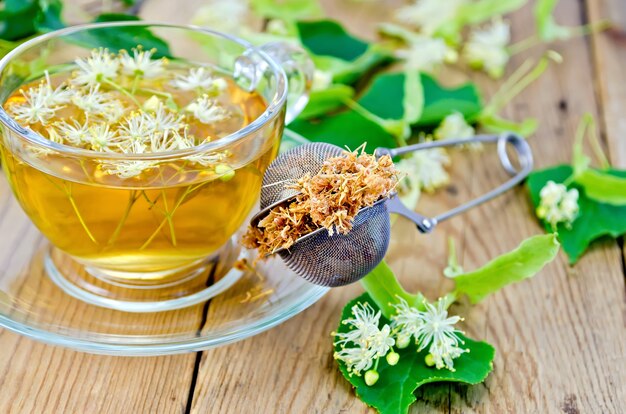 Tamis en métal avec fleurs séchées de tilleul, fleurs fraîches de tilleul, thé dans une tasse en verre sur le des planches en bois