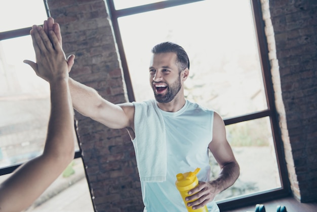 Tape la! Photo de beau mec entraîneur sportif mec s'entraîner à tenir une serviette de bouteille d'eau friendly souriant au client sportswear débardeur formation maison windows à l'intérieur