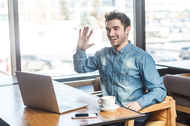 Tape la! Portrait d'un jeune homme d'affaires heureux et satisfait en chemise de jeans bleu assis dans un café et saluant un travailleur via une webcam et montrant la paume de la main avec un sourire à pleines dents.