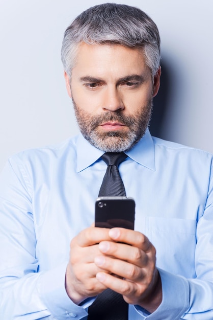 Taper un message commercial. Homme mûr confiant en chemise et cravate tenant un téléphone portable et le regardant en se tenant debout sur fond gris