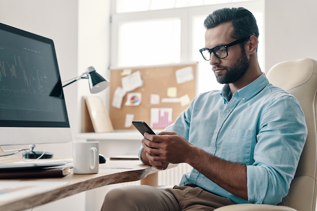 Taper un message commercial. Jeune homme d'affaires moderne utilisant un téléphone intelligent alors qu'il était assis au bureau