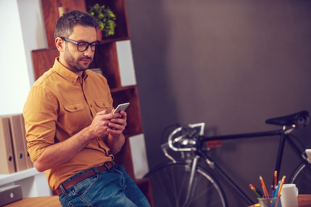 Taper un message commercial. Jeune homme confiant regardant son téléphone intelligent tout en se penchant sur le bureau au bureau