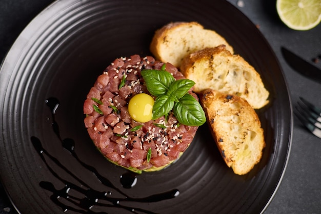 Tartare au thon et à l'avocat avec des graines de sésame, des capers et du jaune d'œuf sur une assiette en céramique foncée avec une baguette