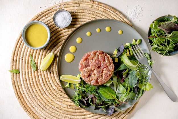 Tartare de thon avec salade verte, citron vert, avocat et sauce moutarde servant sur une plaque en céramique sur une serviette en paille sur fond de texture blanche. Mise à plat, copiez l'espace.