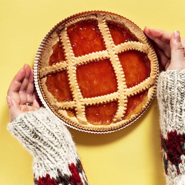 Tarte crostat italienne avec confiture d'abricot sur fond jaune et mains féminines dans un pull en laine chaud avec un ornement