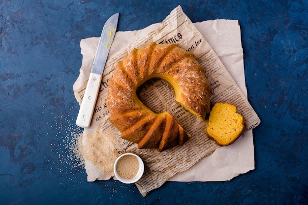 Photo tarte ronde à la citrouille, sur papier kraft, couteau, saucière, cassonade, morceau de gâteau, vue de dessus, fond bleu foncé