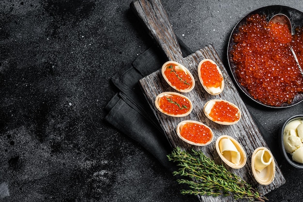 Tartelettes au caviar rouge, beurre et thym sur planche de bois. Fond noir. Vue de dessus. Espace de copie.