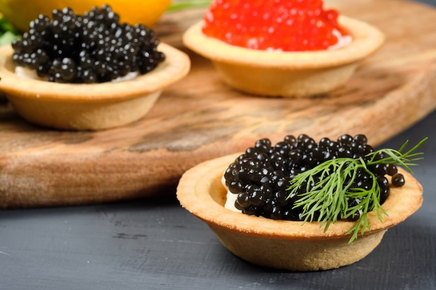 Tartelettes au caviar rouge et noir sur une planche de bois avec du citron et des herbes.