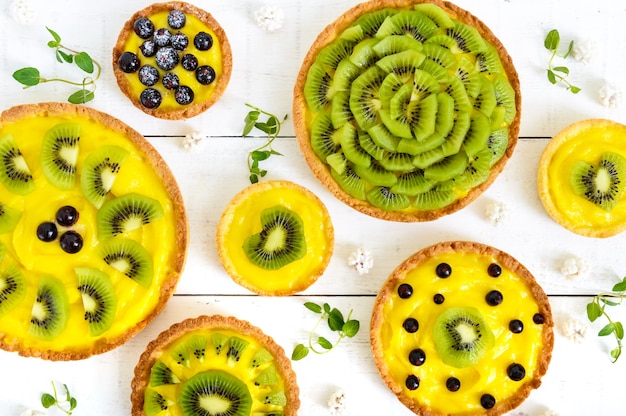 Tartelettes, tartas de crème anglaise, kiwi et cassis de différentes tailles sur une table en bois blanc. Vue de dessus.