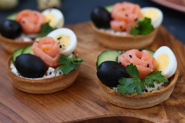 Tartelettes de vacances au fromage, saumon, olives noires, œufs de caille et concombres sur planche de bois sur fond sombre, orientation horizontale, gros plan