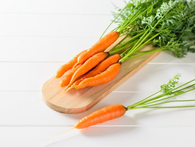 Un tas de carottes fraîches sur une planche à découper et une carotte sur une table en bois blanc.