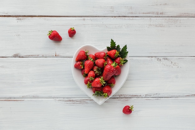 Photo tas de fraises fraîches dans un bol en céramique en forme de coeur sur fond en bois blanc