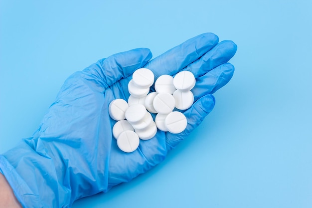 Un tas de pilules se trouve dans une main féminine dans un gant médical sur fond bleu. Soutien sanitaire.