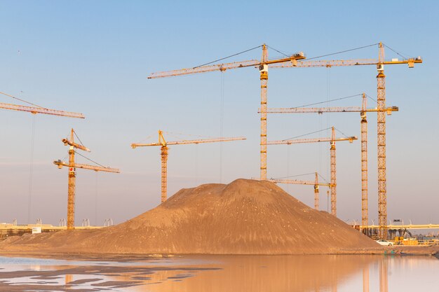 Tas de sable élevé et grues à tour de groupe à haute altitude au chantier de construction, fond de ciel coucher de soleil