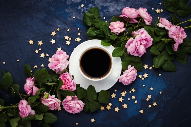 Tasse blanche avec café noir, étoiles et roses roses sur une surface bleu foncé