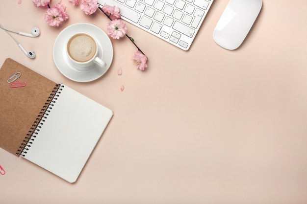 Tasse blanche avec cappuccino, fleurs de sakura, clavier, réveil, bloc-notes sur fond rose pastel