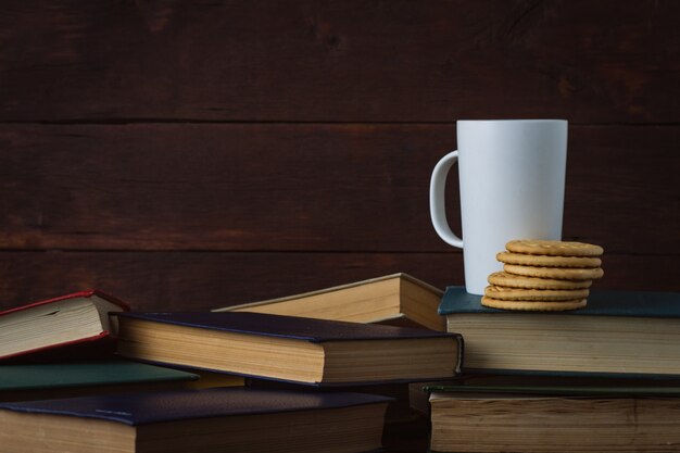 Tasse blanche avec du café chaud, des cookies sur les livres déployés sur un fond en bois foncé