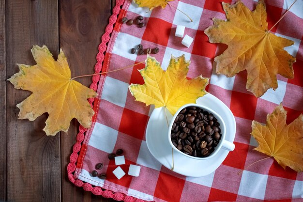 Tasse blanche avec des grains de café sur un fond en bois et une nappe avec des cubes de sucre de cellules rouges et blanches et des feuilles d'automne