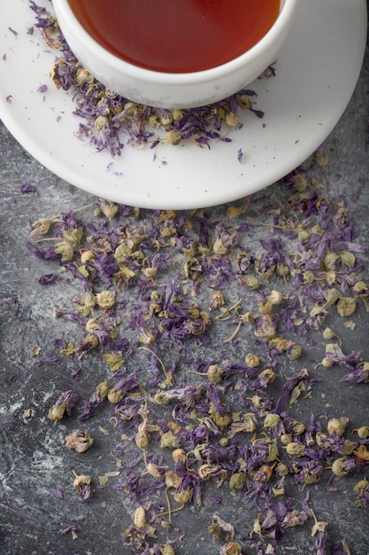 Tasse blanche de thé noir chaud avec des boutons de fleurs séchées placés sur une table gris foncé.