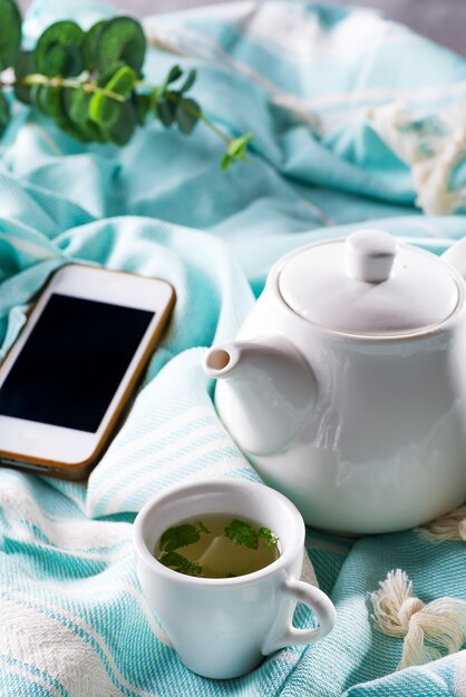 Tasse blanche et théière avec plaid bleu et téléphone sur un lit