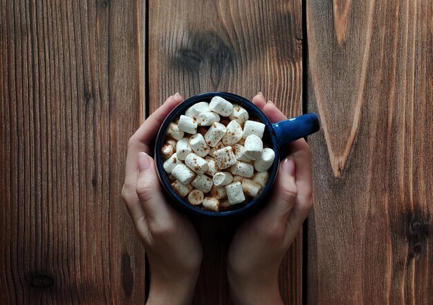 Tasse bleue de chocolat chaud avec des guimauves et de la cannelle dans les mains de la femme, fond en bois, vue de dessus