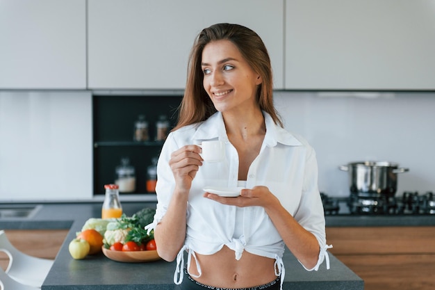 Tasse avec boisson et biscuit Jeune femme européenne est à l'intérieur à la cuisine à l'intérieur avec des aliments sains