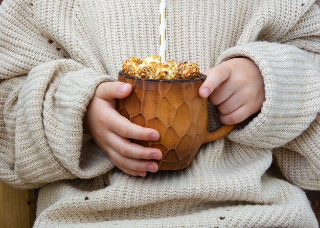 Une tasse avec une boisson chaude au cacao et du pop-corn entre les mains d'un enfant. Maison confortable d'automne, pull chaud
