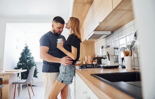 Avec une tasse de boisson fraîche. Matin pour jeune couple marié qui se tient à l'intérieur dans la cuisine.