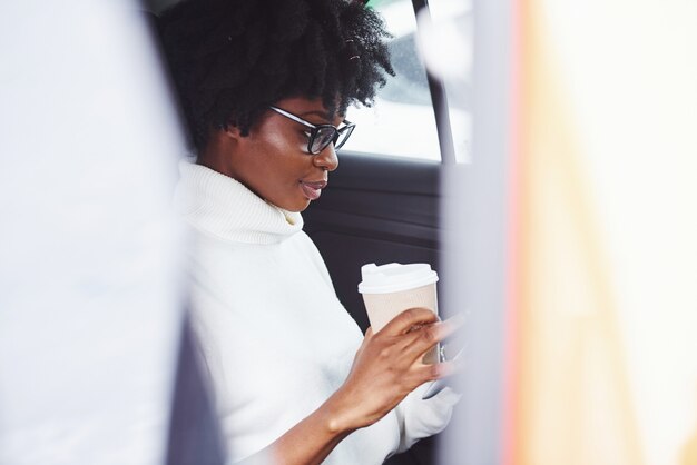 Avec une tasse de boisson. Une jeune femme afro-américaine est assise à l'intérieur d'une nouvelle voiture moderne.