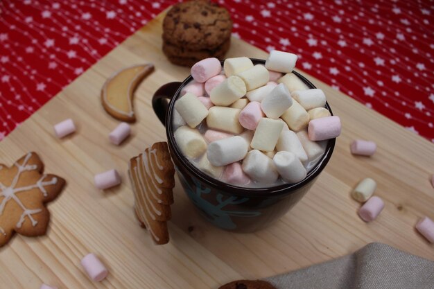 Une tasse brune avec des marshmallows, des biscuits avec des formes de Noël, un chiffon de cuisine gris, du bois et une nappe rouge.