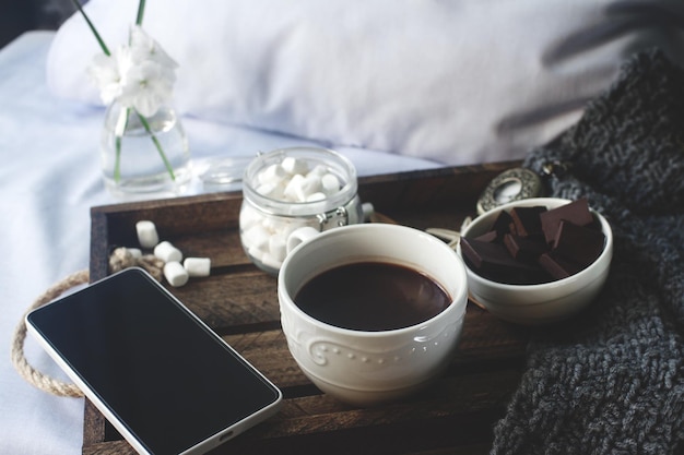 Tasse de cacao maison avec des fleurs de chocolat à la guimauve et un smartphone sur un plateau en bois rustique dans le lit douillet week-end matin blogueur style de vie mise au point sélective