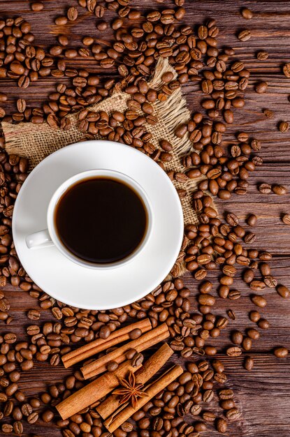 Une tasse de café aromatique noir dans une tasse blanche avec des grains de café torréfiés arabica, des étoiles d'anis, des bâtons de cannelle sur un fond en bois