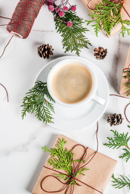 Tasse à café au lait avec cadeau de Noël ou boîte cadeau enveloppée dans du papier kraft, décorée de branches d'arbres de Noël, pommes de pin, fruits rouges, sur une table en marbre blanc, vue de dessus