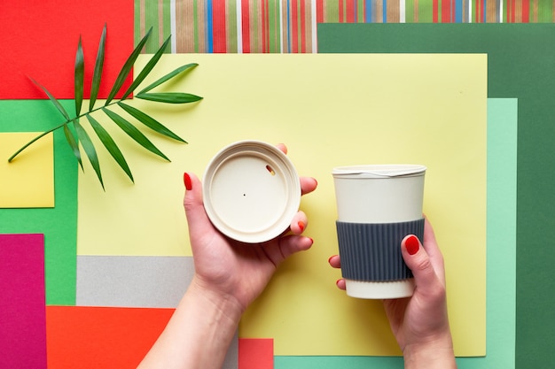 Tasse à café en bambou avec couvercle en mains féminines sur fond de papier rectangle multicolore géométrique abstrait avec feuille de palmier. Mise à plat créative avec tasse biodégradable zéro déchet.