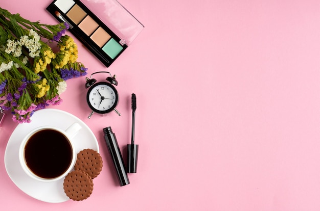 Tasse à café avec des biscuits, réveil, fleurs, mascara, sur une surface rose.