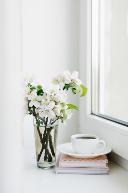 Tasse de café blanche sur une assiette blanche une pile de livres et un vase de fleurs sur le rebord de la fenêtre Cosy Easter spring still life