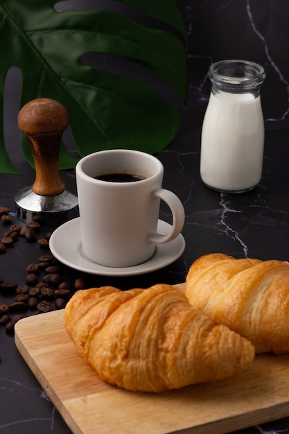 Une tasse de café blanche a été placée à côté d'une bouteille de lait et un croissant sur une planche à découper, des grains de café et des moulins sur un sol en marbre.