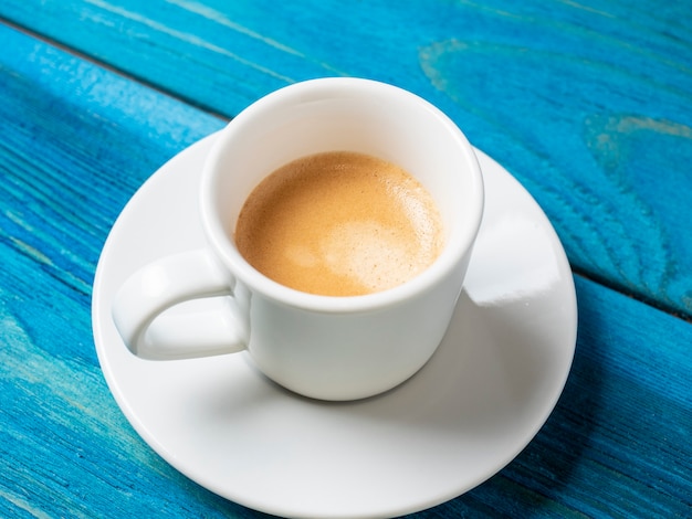 Une tasse à café blanche avec un expresso complet est posée sur une soucoupe et sur un fond en bois bleu