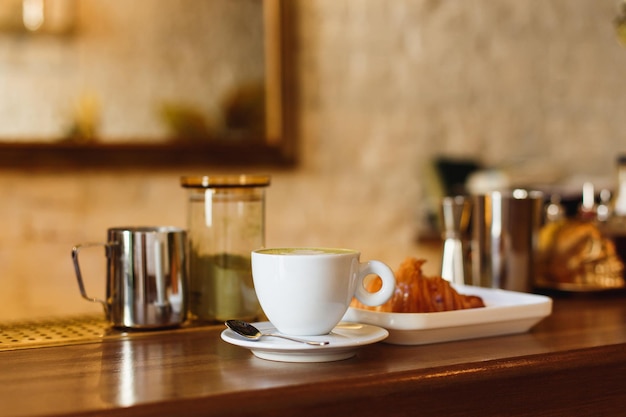 Tasse à café blanche avec matcha cappucsino et une assiette avec un croissant sur un comptoir de bar en bois