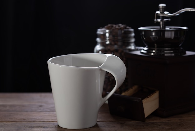 Tasse à café blanche, moulin à café et grains torréfiés sur table en bois avec fond sombre