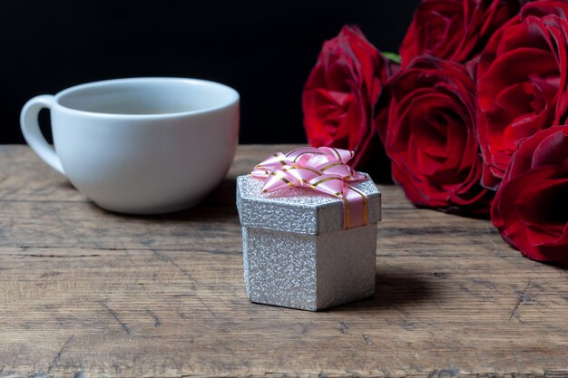 Tasse à café, boîte-cadeau avec ruban rose et roses rouges derrière.