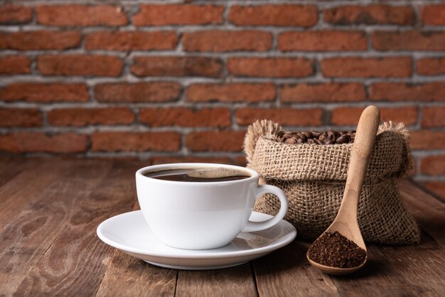 Tasse à café, café moulu et grains de café en toile de jute sur table en bois avec mur de briques