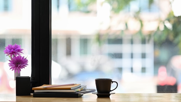 Tasse à café en céramique noire avec pile de cahier et plante en pot sur la table à bois moderne avec fenêtres noires recadrées